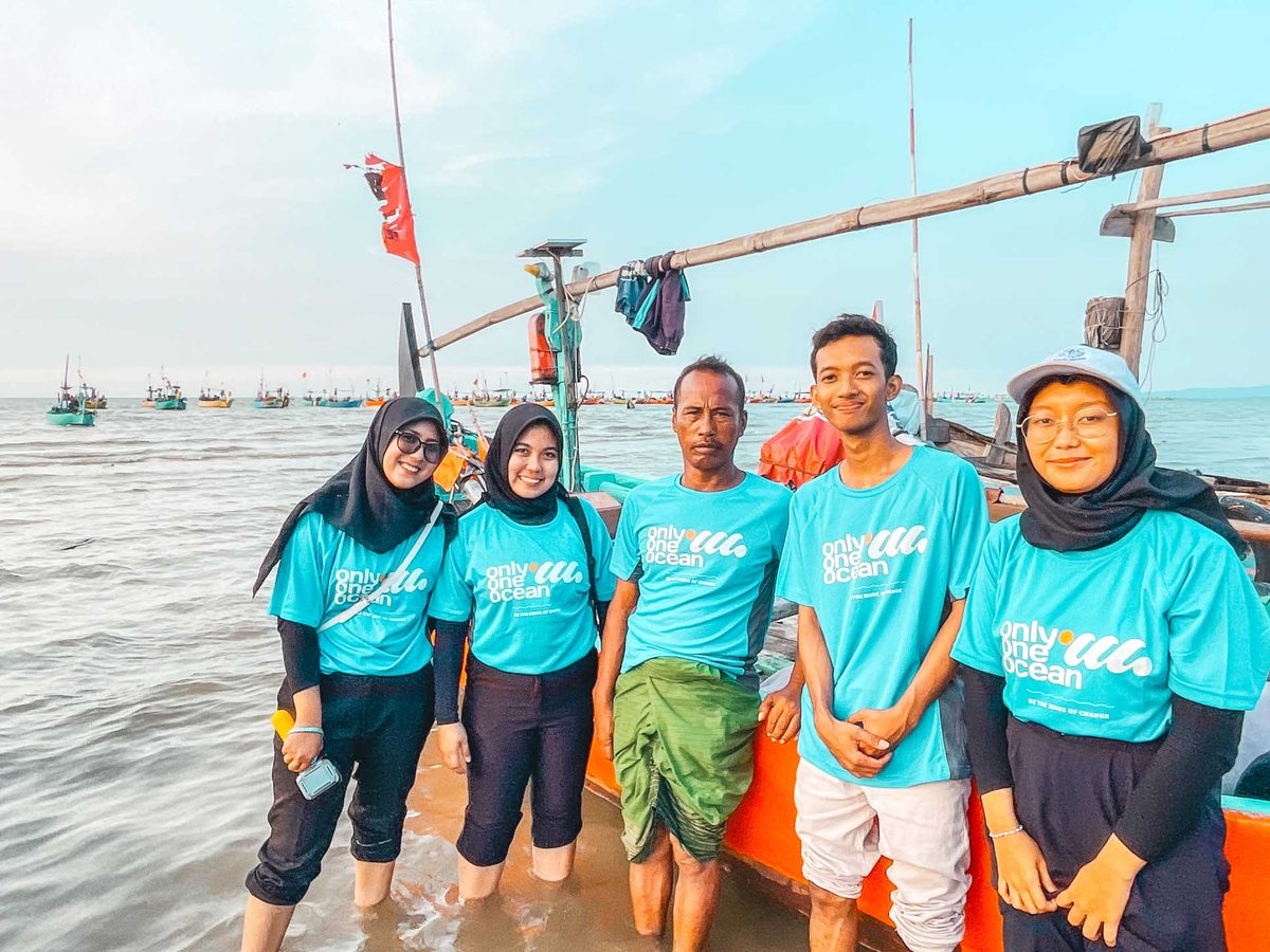 Five O3 team members in matching aqua shirts standing on the shore with fishing boats behind them, at the end of a community engagement session.