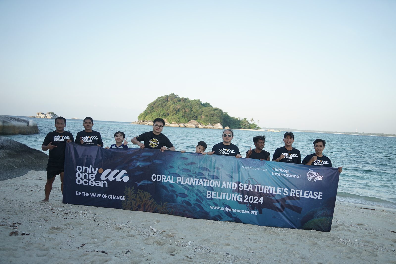 The full O3 field team standing on a Belitung beach holding a banner reading Coral Plantation and Sea Turtle's Release, Belitung 2024. A small granite islet sits in the distance behind them, evening light on the water. Partner logos for FishLog International and SeaTracer visible on the banner.